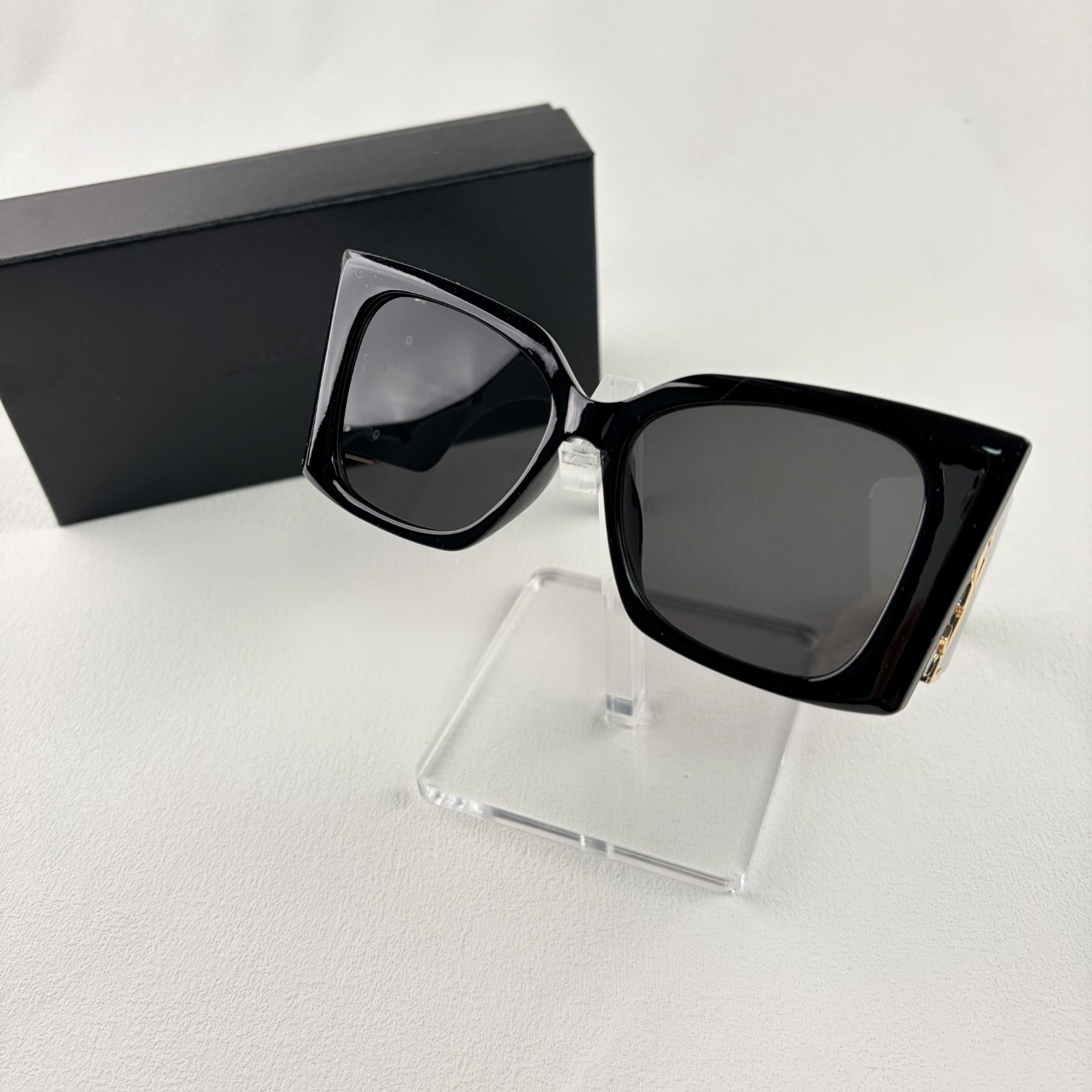 Pair of oversized black square sunglasses with gold metal logo accents on temples on display stand.
