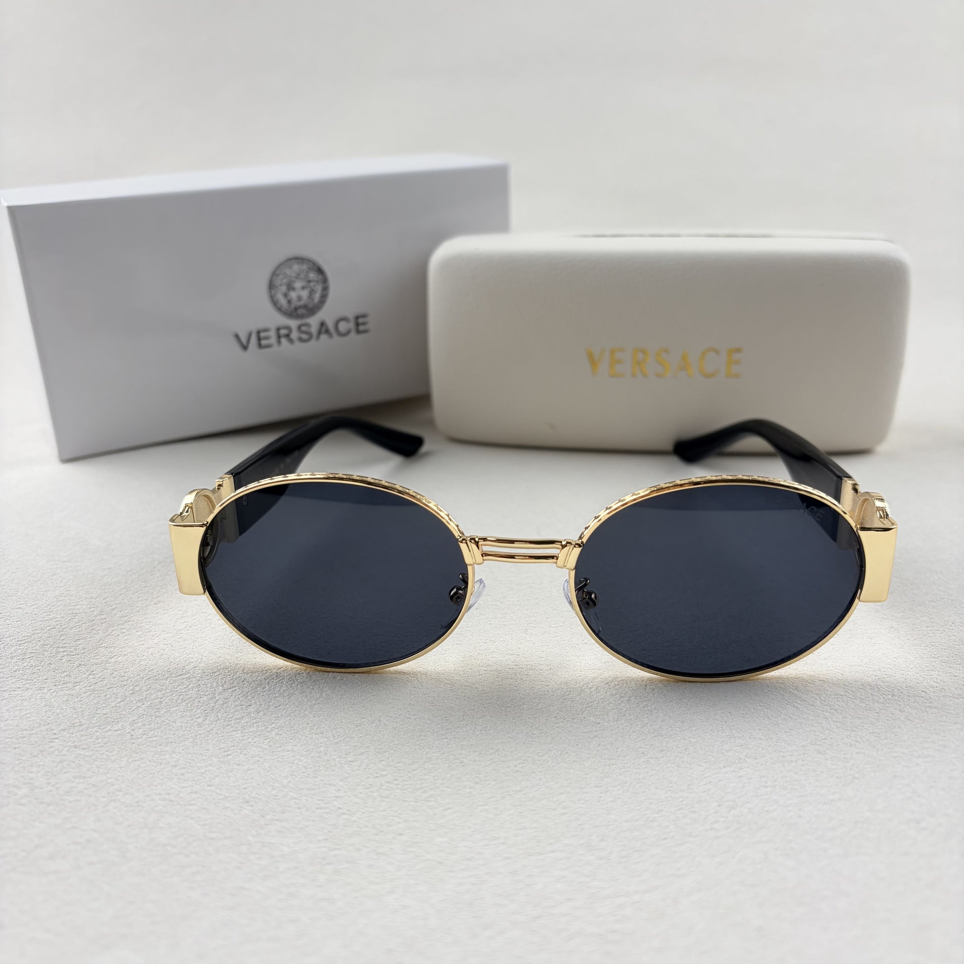 Oval gold metal frame sunglasses with black temples and dark tinted lenses, shown with a storage cas
