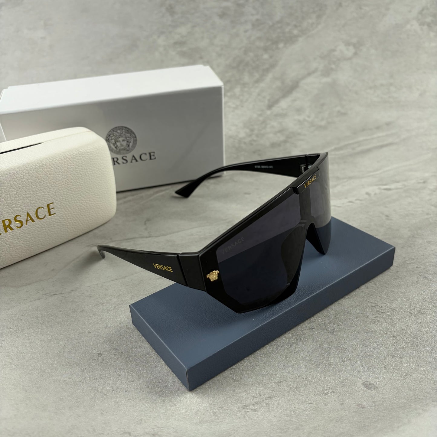 Oversized black shield sunglasses with gold metal accents on arms resting on a blue display box.