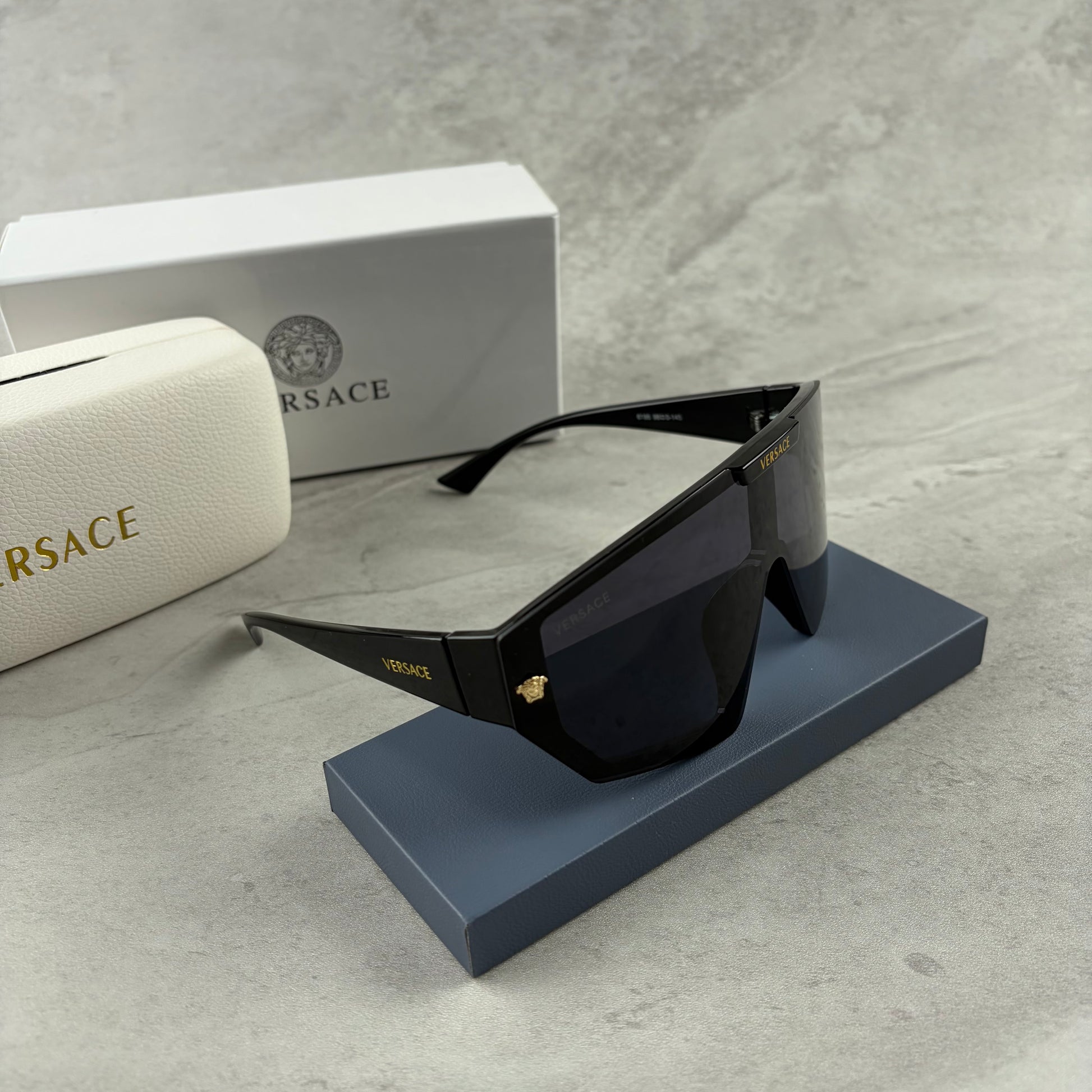 Oversized black shield sunglasses with gold metal accents on arms resting on a blue display box.