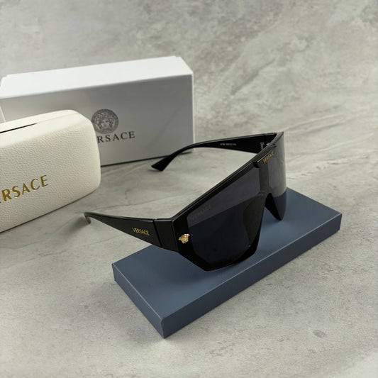 Oversized black shield sunglasses with gold metal accents on arms resting on a blue display box.