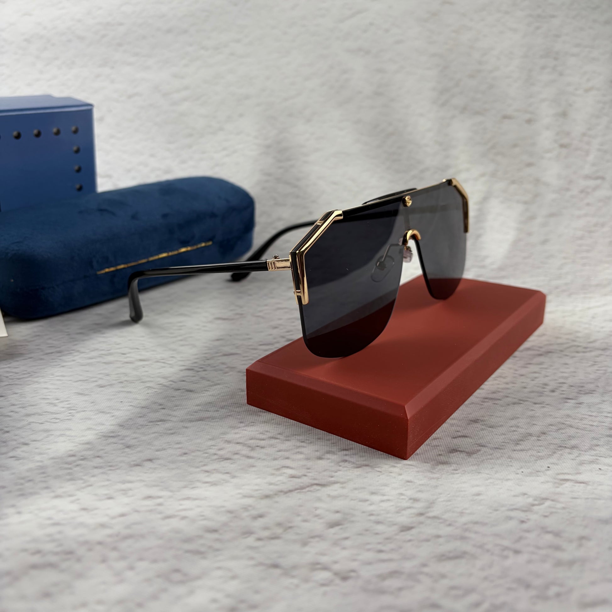 Oversized shield sunglasses with a gold metal frame and dark black lenses on a red display stand.