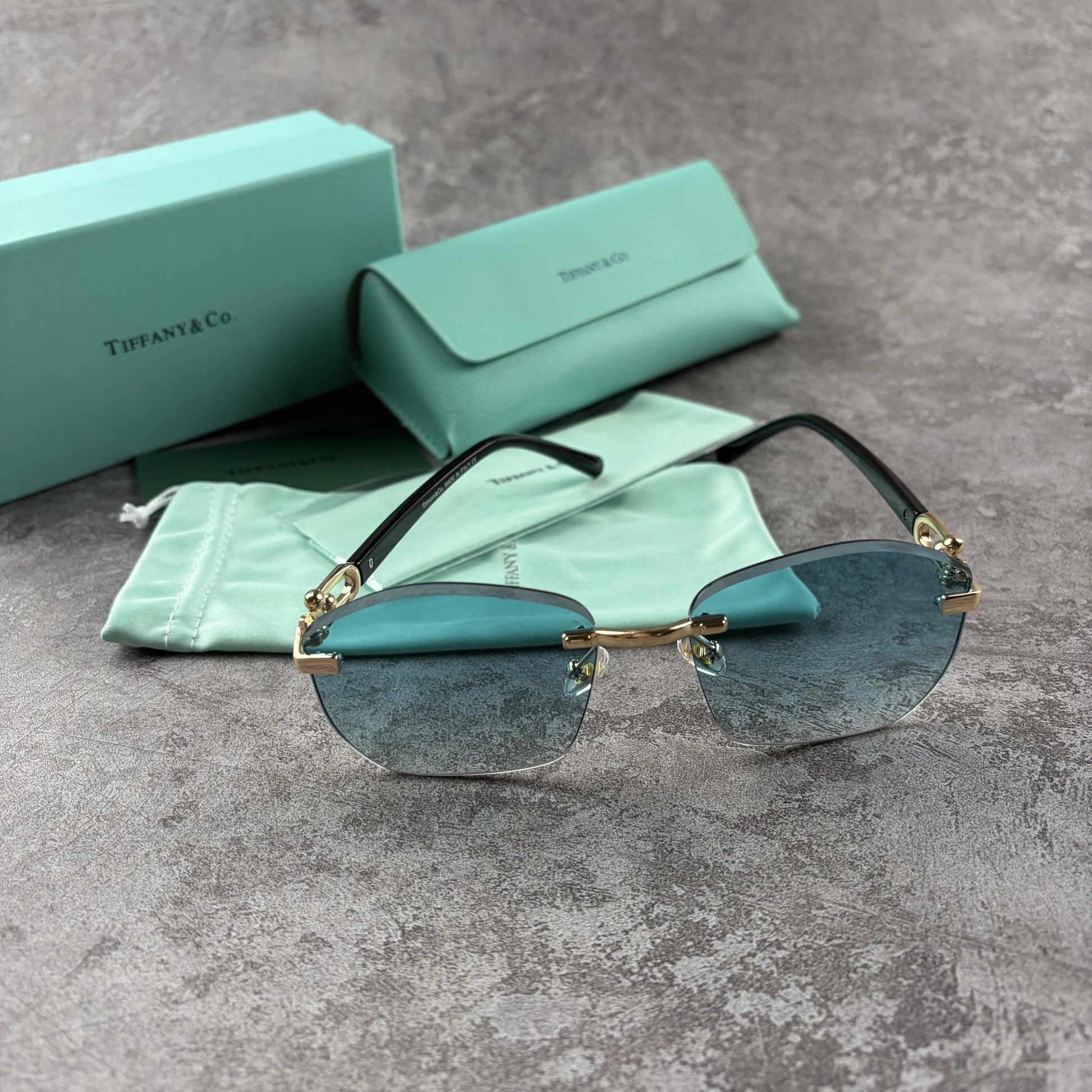 Rimless sunglasses with gradient blue lenses and gold-tone metal arms, displayed with protective cas