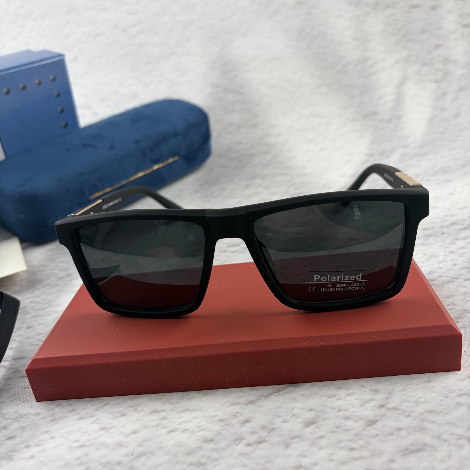 Matte black polarized sunglasses with gold logo detailing on temple arms, sitting on a red display b